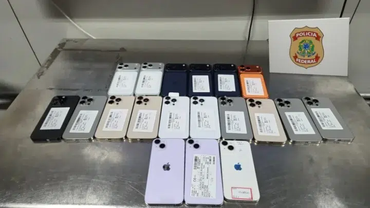 Vários celulares e aparelhos de telefone em diferentes cores, dispostos lado a lado em uma superfície metálica, com etiquetas e alguns com a marcações de identificação em papel.