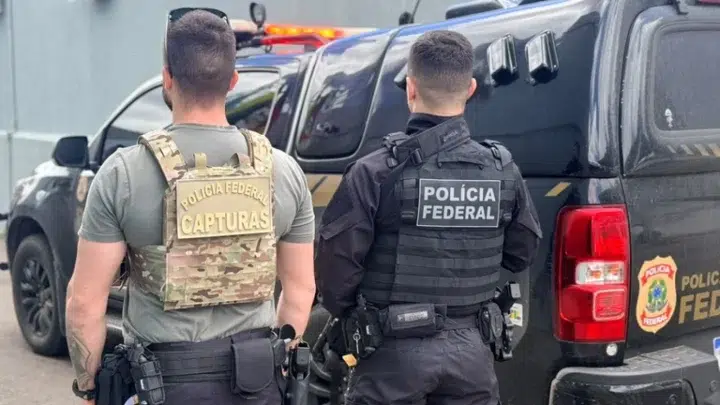 Policiais federais com coletes e mochilas táticas permanecem ao lado de uma viatura estacionada, com o compartimento traseiro aberto.