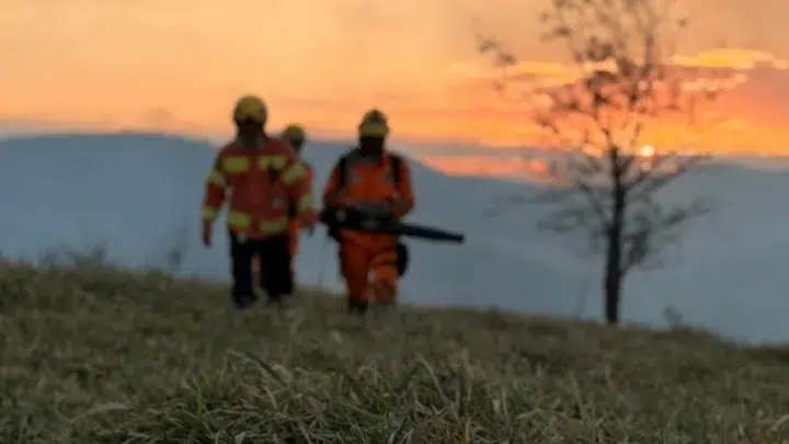 Dois bombeiros caminham por uma área de vegetação em um terreno aberto ao entardecer, com o céu alaranjado ao fundo.