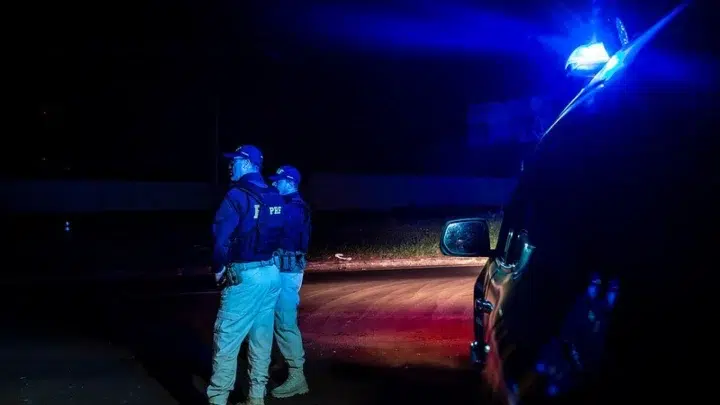 Dois agentes da PRF em destaque, com coletes refletivos azuis, ao lado de uma via noturna iluminada por faróis e luzes de emergência, com veículos envolvidos ao fundo após uma colisão.