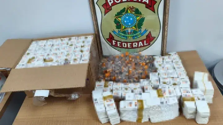 Caixas de papelão e pacotes com frascos/ampolas brancas sobre uma mesa, com um emblema “Polícia Federal” ao fundo.