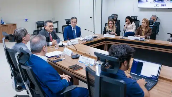 Fotografia da coletiva de imprensa da Operação Jogos de Sombras. Na ponta da mesa estão o subprocurador-geral da República e coordenador do Gaeco Nacional, José Adonis Callou de Araújo Sá, de terno cinza e gravata vermelha fala ao microfone e o secretário especial da Receita Federal do Brasil, Robinson Barreirinhas. Ao lado dele, à mesa mulheres e homens escutam com papéis, cadernos e notebooks à frente. Ao fundo, no canto superior direito, há uma tela de televisão fixada na parede exibindo uma apresentação com as marcas do Ministério Público Federal e da Receita Federal.