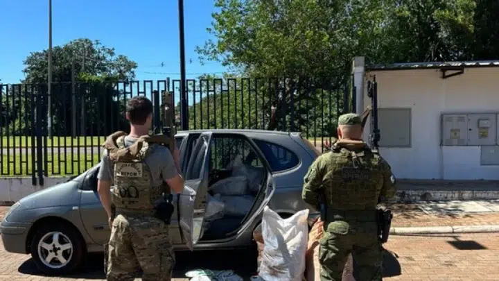 Dois agentes com uniformes operacionais em frente a um carro estacionado e a sacos e pacotes no chão, em área externa pavimentada com portão ao fundo