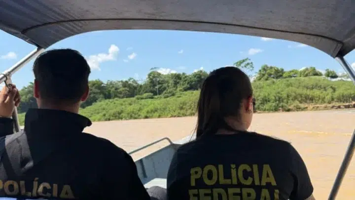 Dois agentes em uma lancha, com camisetas da Polícia Federal, observando a área ao redor enquanto navegam em um rio com vegetação ao fundo.