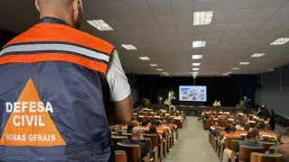 Agente da Defesa Civil com colete laranja e azul, escrito “Defesa Civil Minas Gerais”, em auditório durante um seminário.