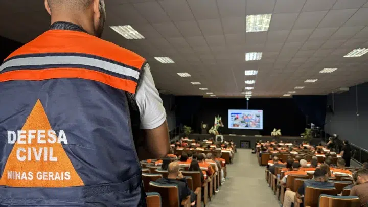 Agente da Defesa Civil com colete laranja e azul, escrito “Defesa Civil Minas Gerais”, em auditório durante um seminário.