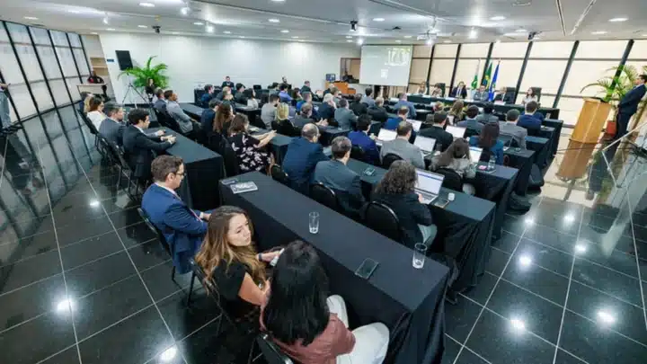 Pessoas em traje formal participam num evento num auditório espaçoso, dispostas em mesas cobertas com toalhas pretas viradas para uma mesa principal com bandeiras e um ecrã de projeção ao fundo.